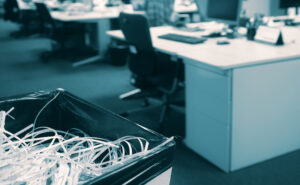 A close-up of a trash bin filled with shredded paper in an office with desks and computers in the background.